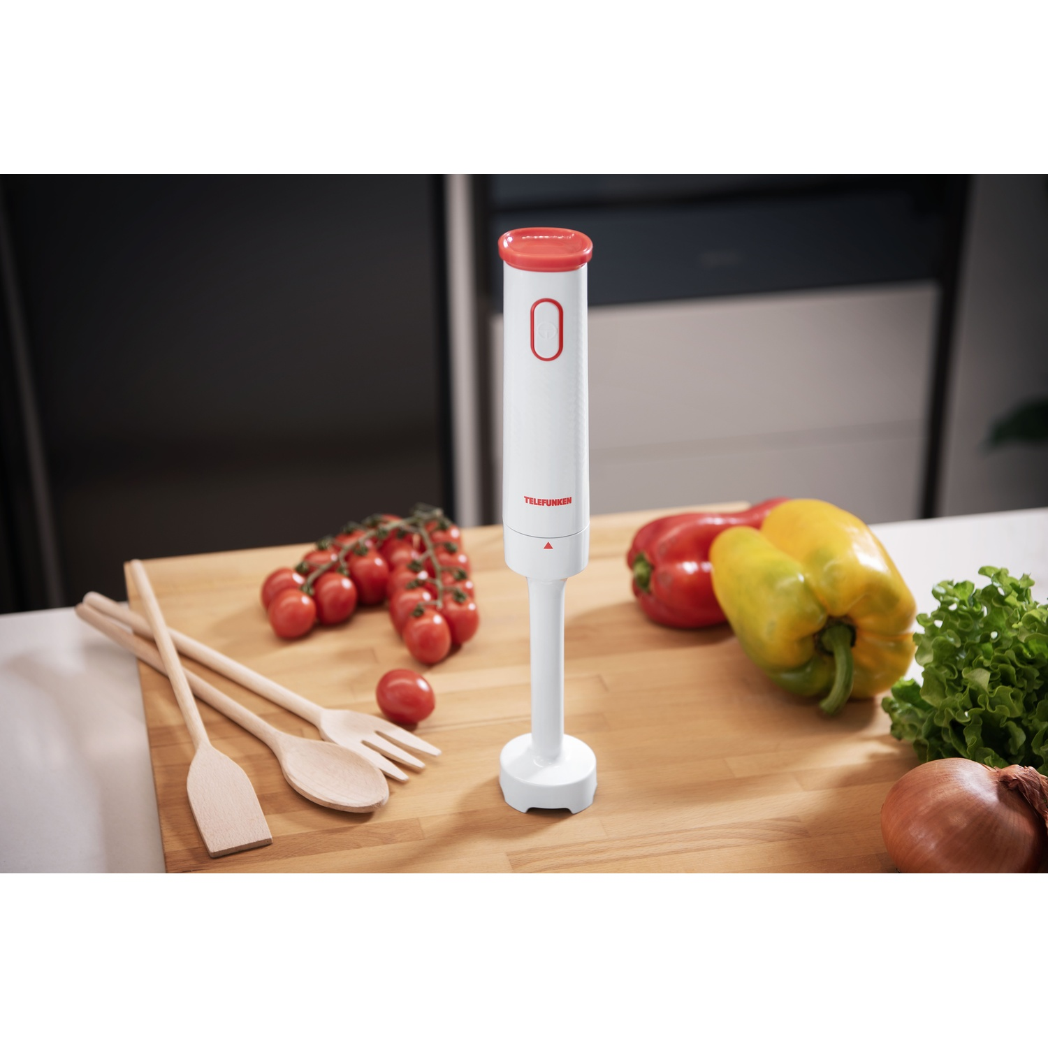Un frullatore a immersione bianco con parte superiore rossa. Pomodori, peperoni, lattuga e cipolla su tagliere di legno.