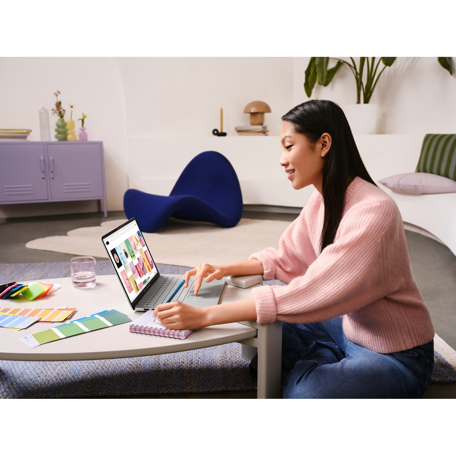 Una donna in maglione rosa usa un laptop, seduta sul pavimento con un taccuino. Tavolozze di colori e un bicchiere d'acqua sono vicini.