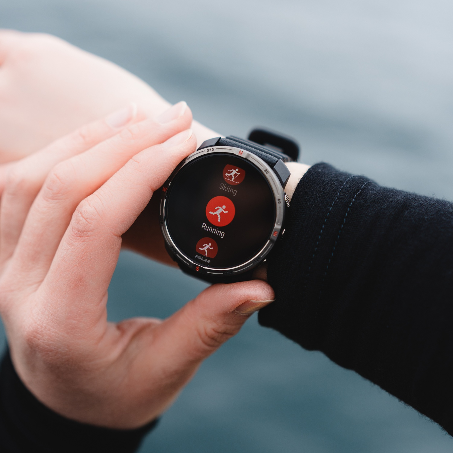 A person wearing a smartwatch displays skiing and running activities. The watch is on a wrist, a dark blue jacket is visible.