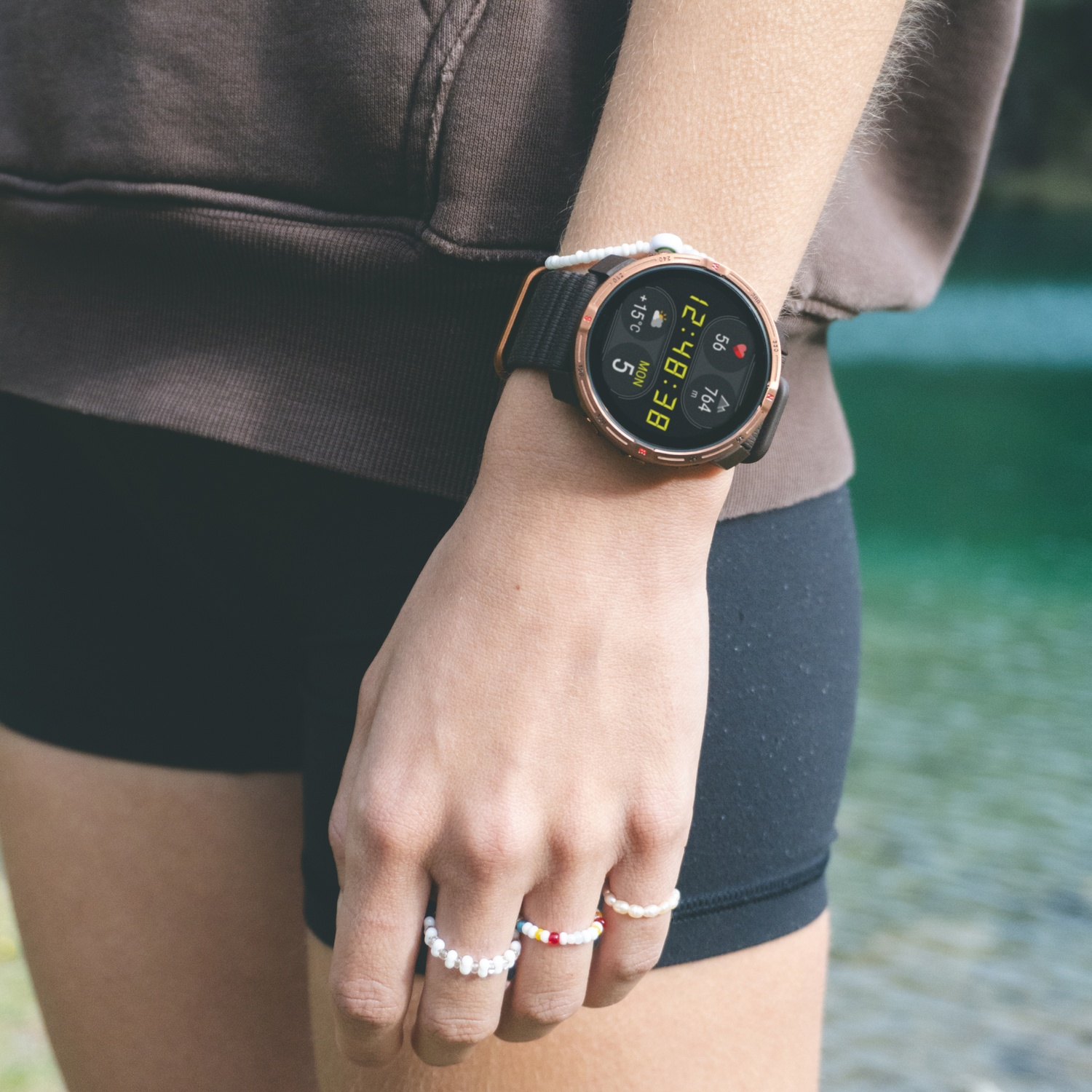 Une personne portant une smartwatch affichant l'heure et les statistiques. Elle porte des bagues, un short et un sweat à capuche marron. Le fond est de l'eau.