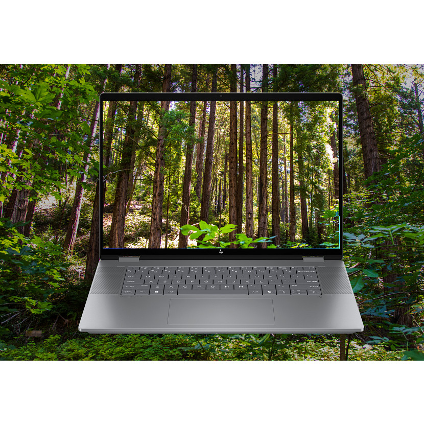 Laptop wyświetlający scenę leśną. Laptop jest srebrny, z klawiaturą. Tło to gęsty zielony las.