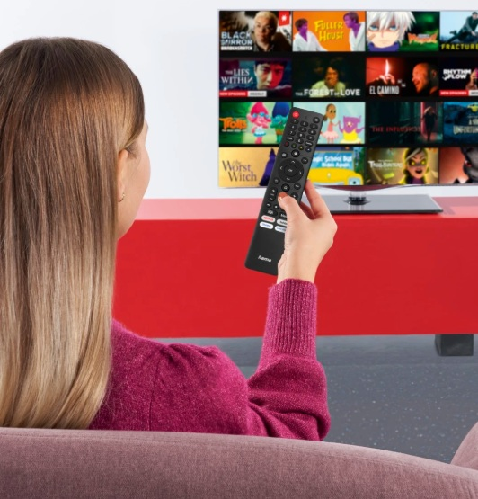 Kobieta używa pilota do nawigacji po telewizorze, na którym wyświetlane są różne tytuły filmów. Ma na sobie różowy sweter.