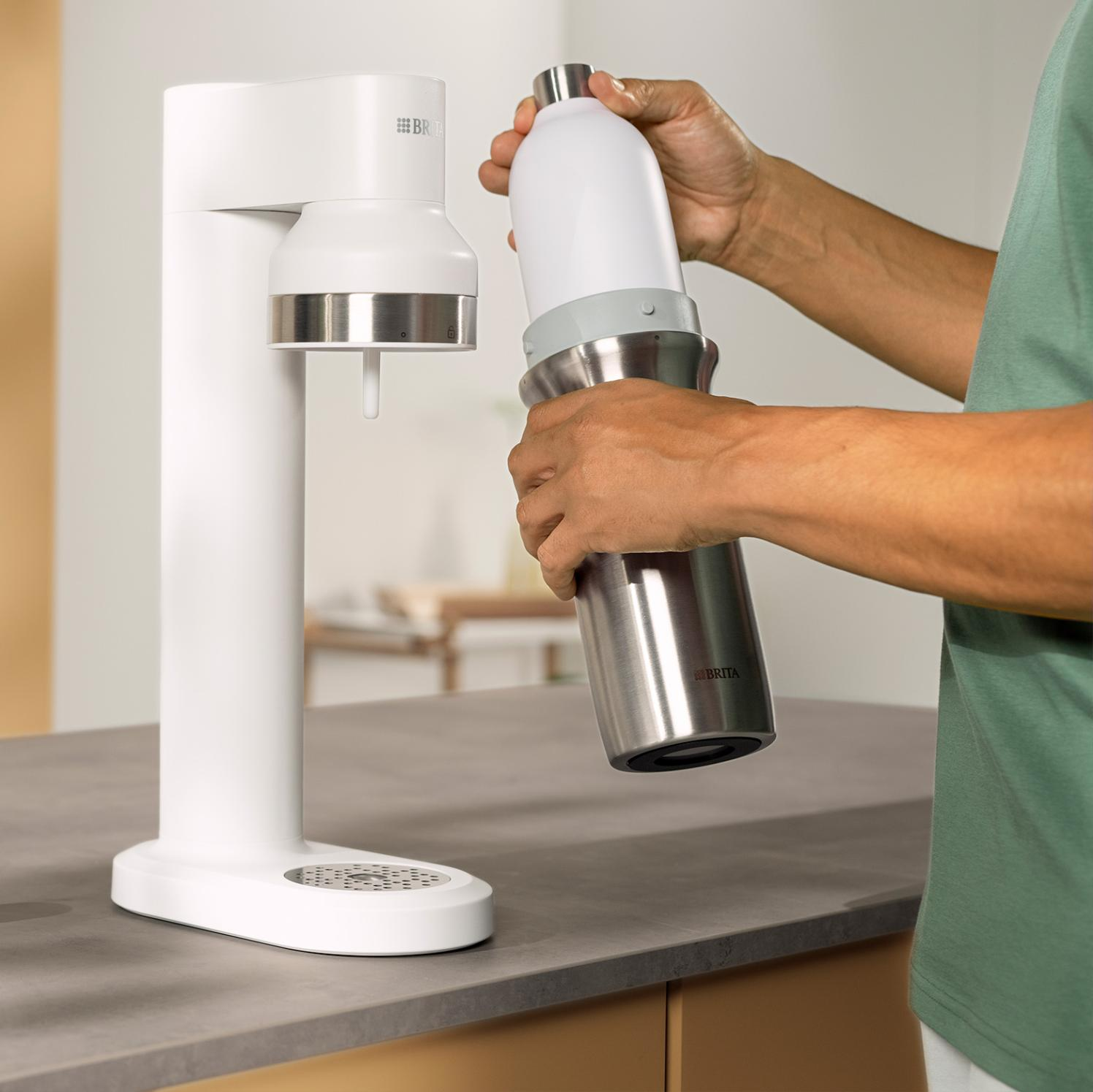 Osoba używa białego saturatora. Mocuje srebrną butelkę do urządzenia. Scena rozgrywa się na blacie kuchennym.