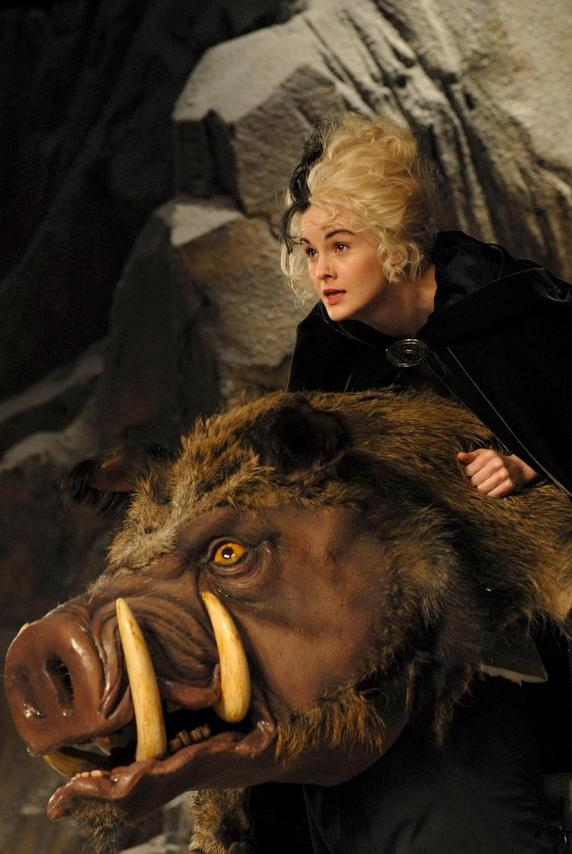 Eine Frau mit schwarz-weißer Frisur steht hinter einem großen Eberkopf. Das Wildschwein hat große Stoßzähne und gelbe Augen. Die Frau trägt Schwarz.