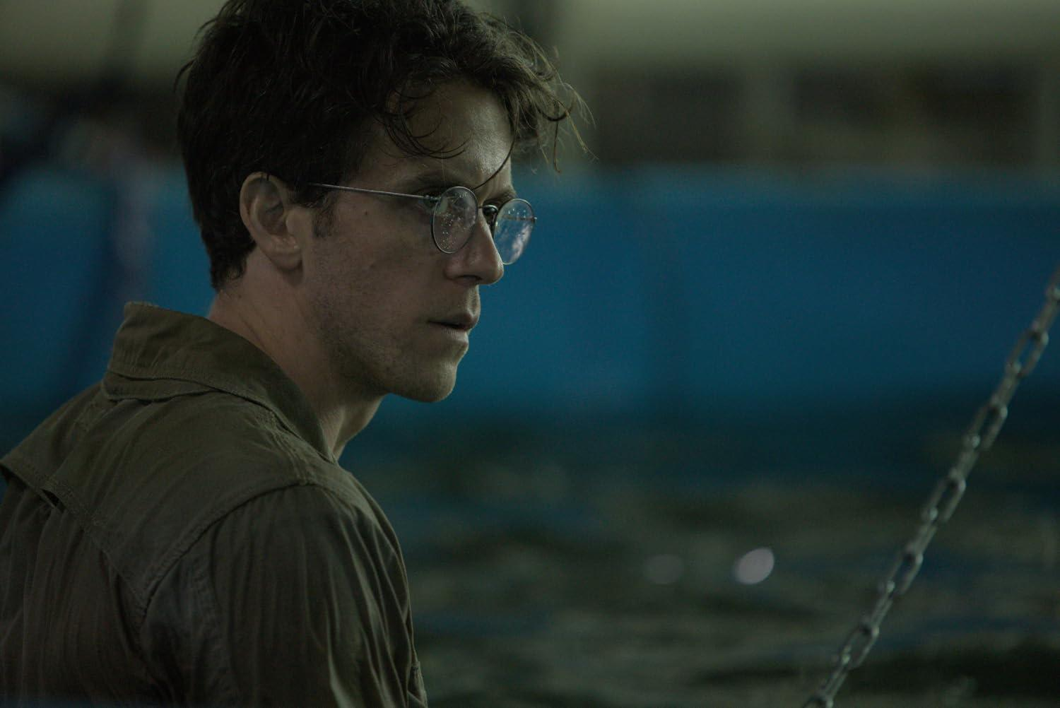 Ein Mann mit Brille und grünem Hemd blickt zur Seite. Der Hintergrund ist blau und verschwommen. Rechts ist eine Kette zu sehen.
