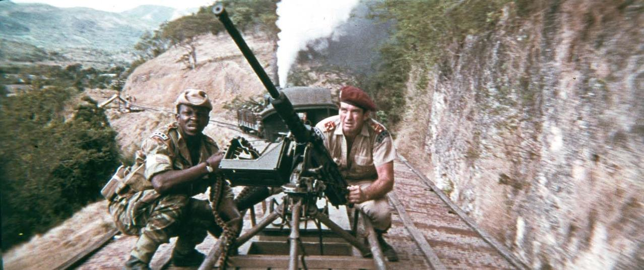 Zwei Soldaten mit einer großen Kanone auf Bahngleisen, Berge im Hintergrund. Einer trägt Barett, der andere eine Kappe.