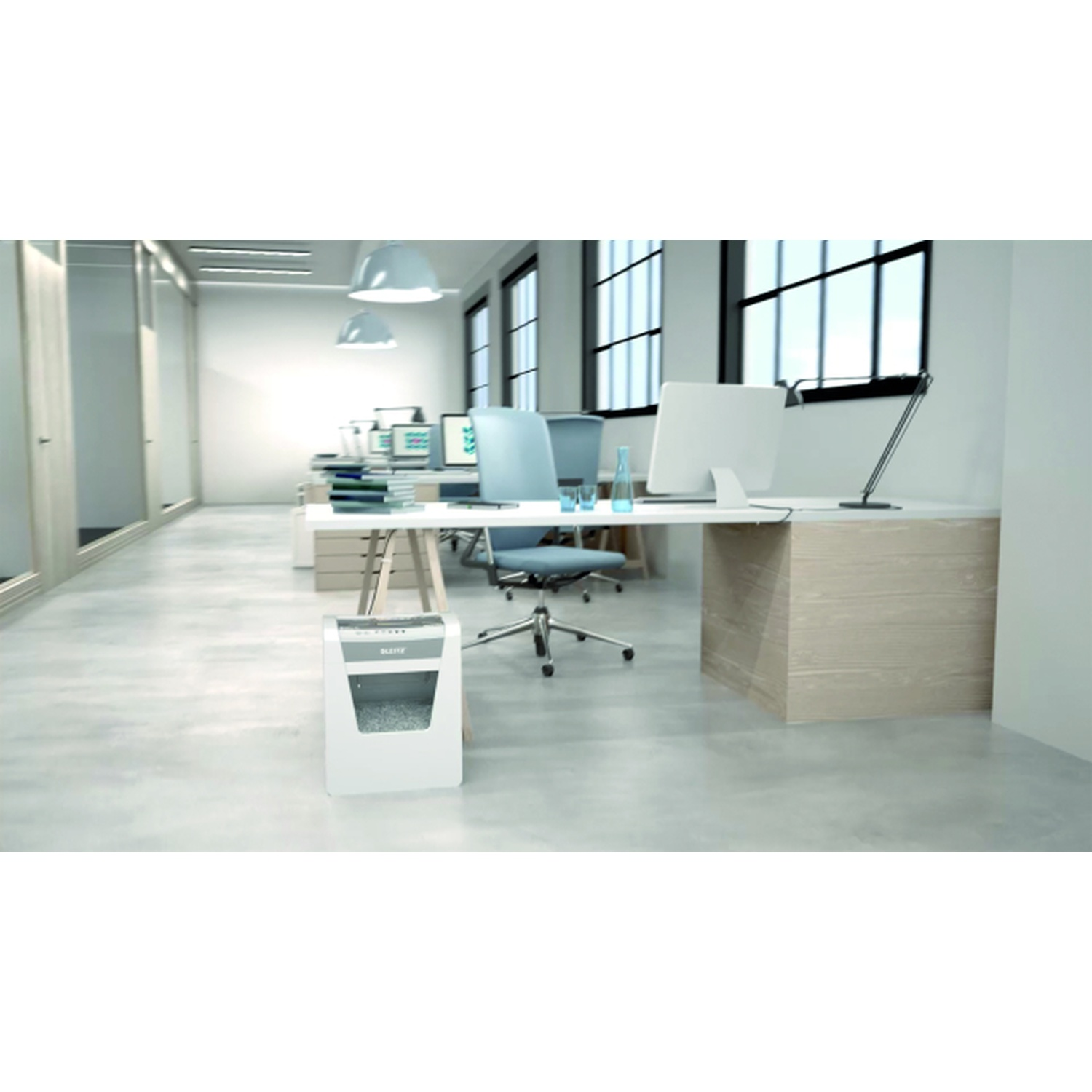 Un ufficio con scrivanie, sedie, computer e un tritadocumenti. Finestre sullo sfondo. Spazio di lavoro moderno e ben illuminato.