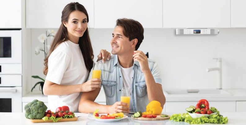 Para w kuchni uśmiecha się, trzymając szklanki soku. Warzywa leżą na stole. Nad nimi znajduje się wieża stereo.