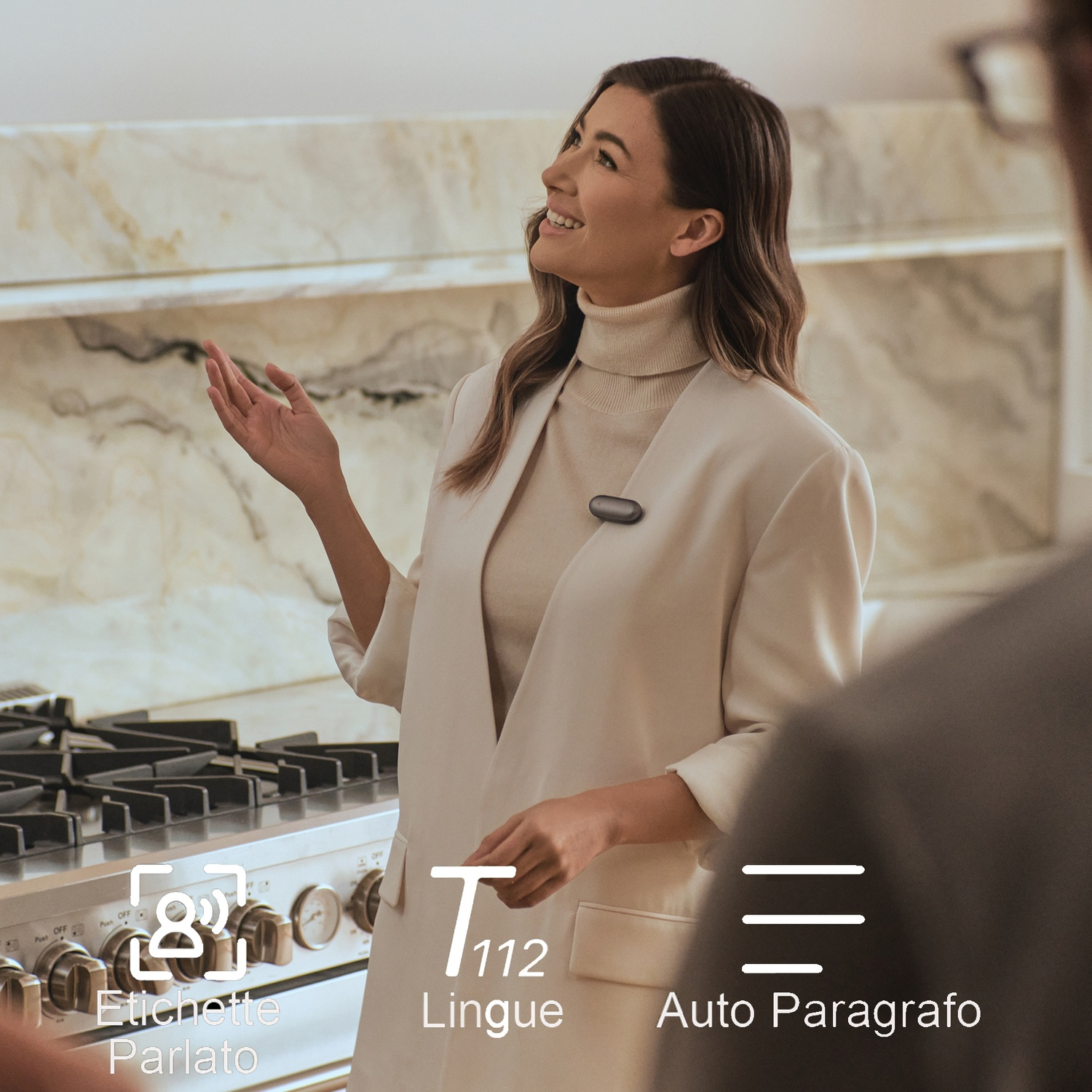 Una donna gesticola mentre parla. Indossa una giacca bianca e un dolcevita beige. Elettrodomestici da cucina sullo sfondo.