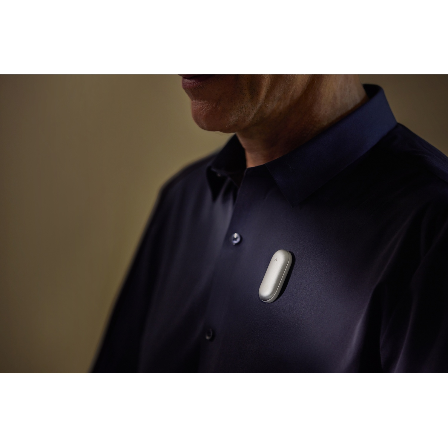 Un uomo indossa una camicia blu navy, con un piccolo dispositivo argentato attaccato. Lo sfondo è color cachi sfocato.