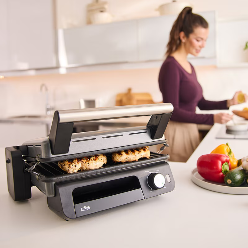 Grill csirkével. A háttérben egy nő. Zöldségek vannak az asztalon.