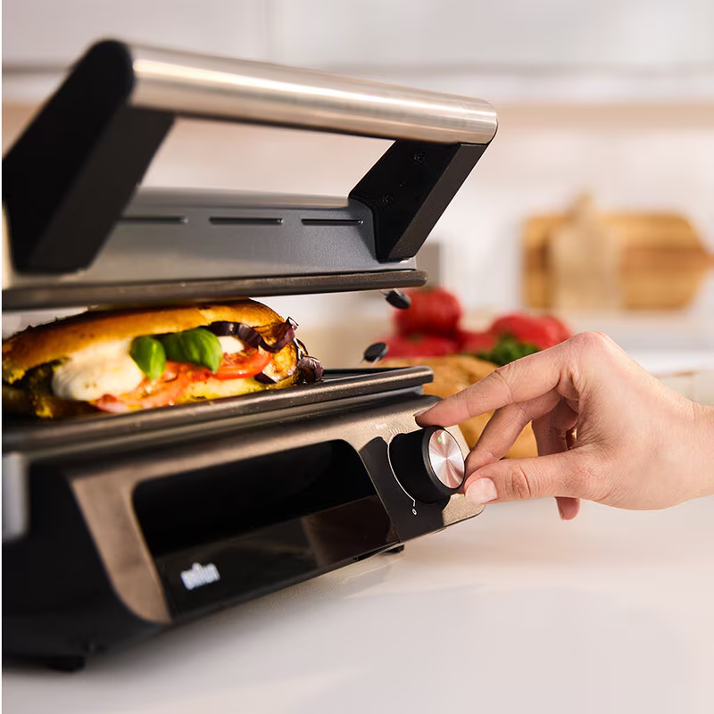Egy személy beállítja a szendvicsgrillt. Egy szendvics paradicsommal, mozzarellával és bazsalikommal van benne. Egyéb hozzávalók a háttérben.