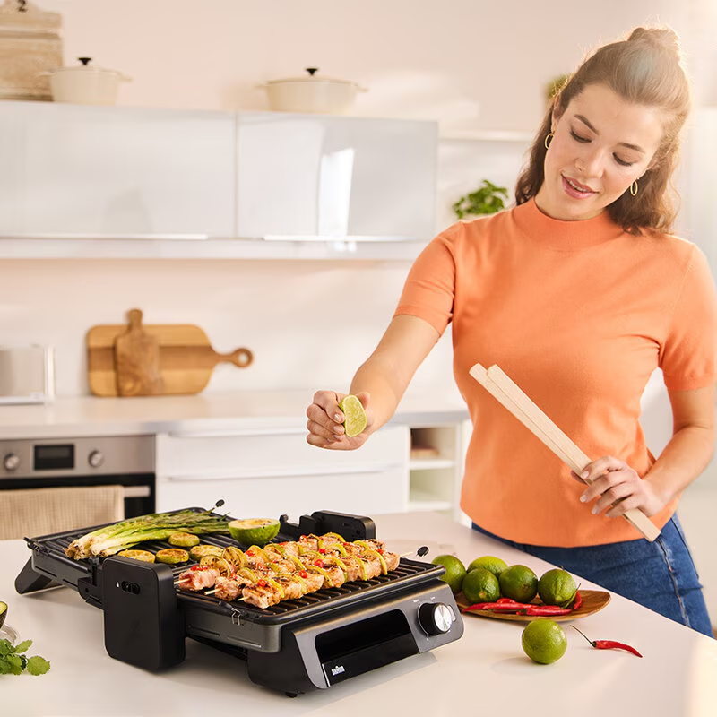 Egy nő limelevet nyom a grillezett nyársakra. Lime, grill és zöldségek vannak a pulton. A háttérben egy konyha látható.