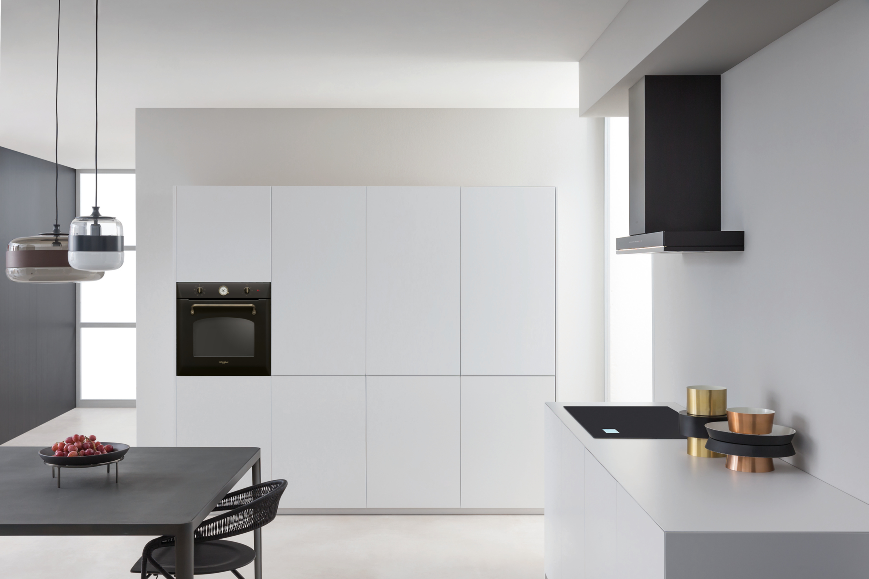 Nowoczesna kuchnia z białymi szafkami, czarnym piekarnikiem i ciemnym stołem z owocami. Wiszące lampy.