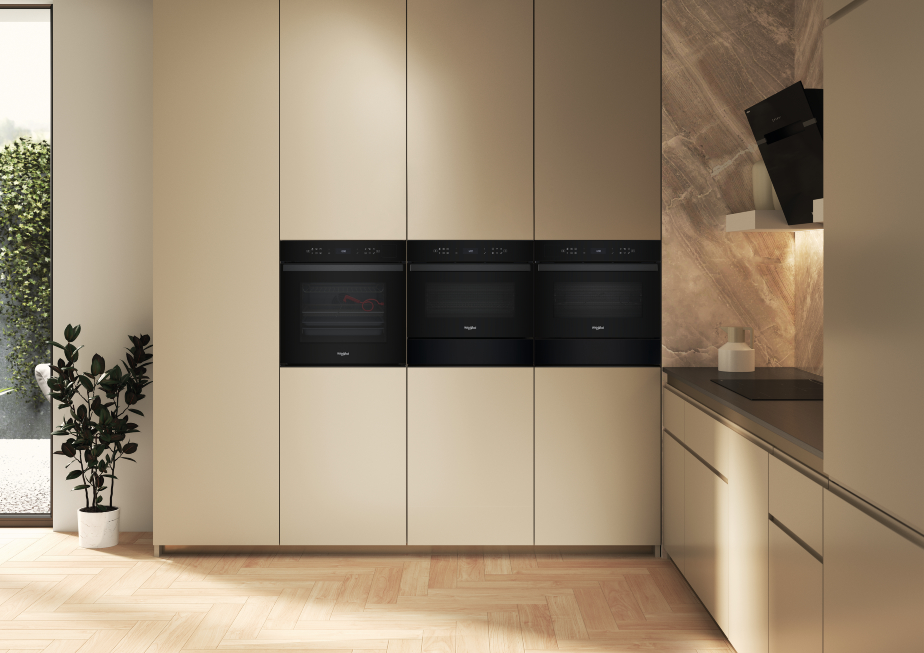 Wnętrze kuchni z trzema czarnymi piekarnikami, drewnianą podłogą i rośliną w białej doniczce. Jasnobeżowe szafki.