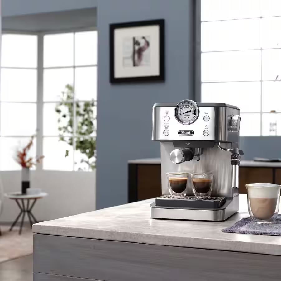 Ezüst eszpresszógép két eszpresszó pohárral. Jobbra egy latte pohár. Konyhai környezet, háttérben ablakokkal.