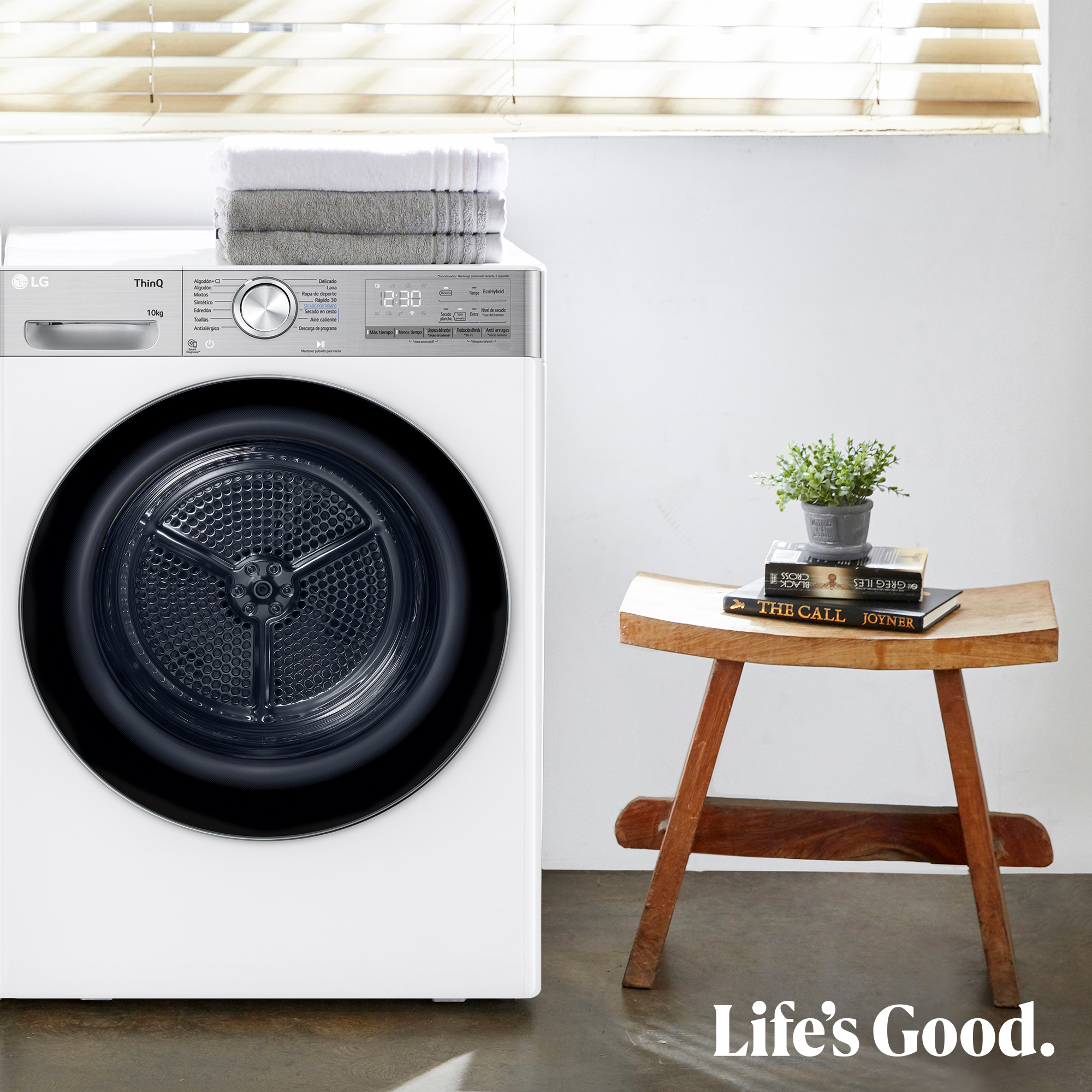 Una secadora LG blanca con toallas encima, un taburete de madera con libros y una planta y el texto 'Life's Good'.