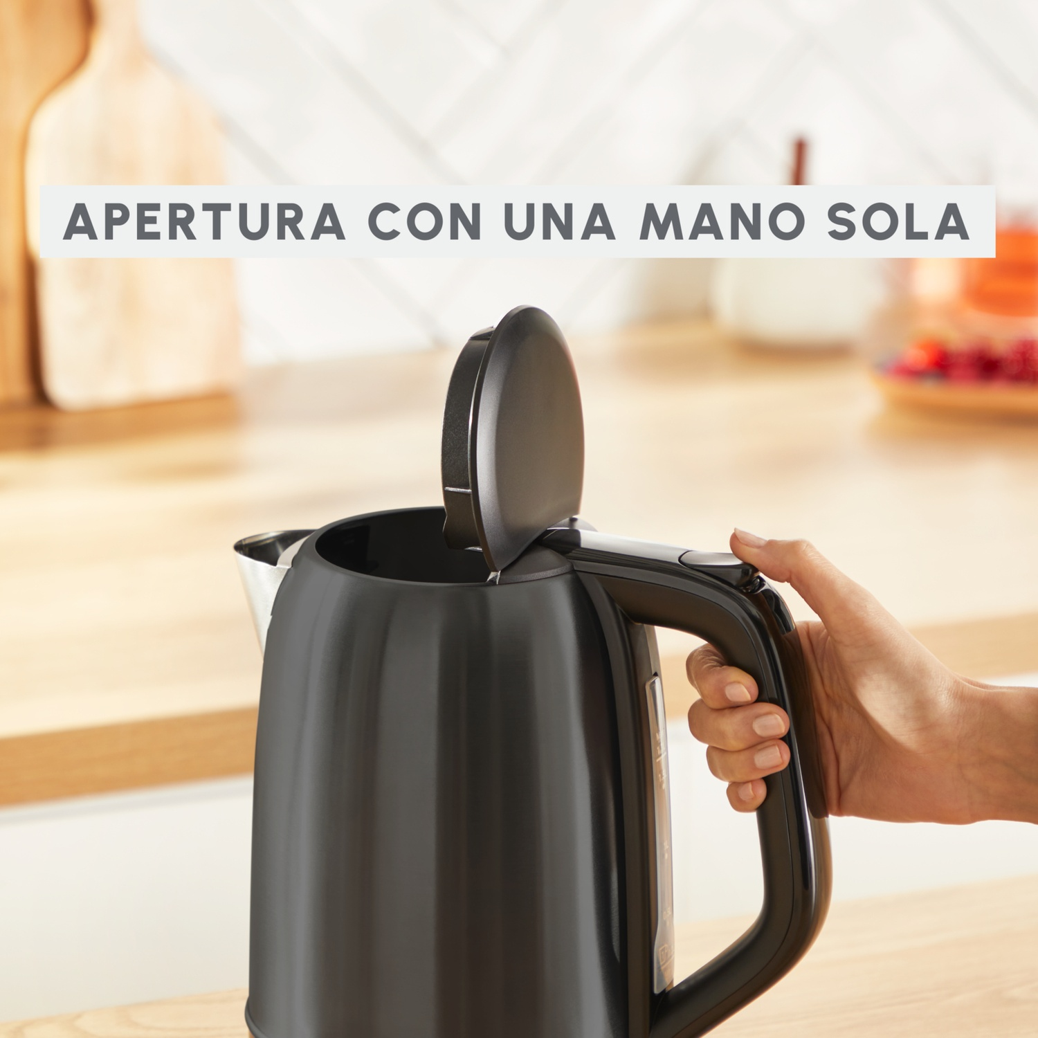 Un bollitore elettrico nero viene aperto con una mano. Il coperchio è sollevato. Lo sfondo comprende una superficie in legno e altri oggetti.