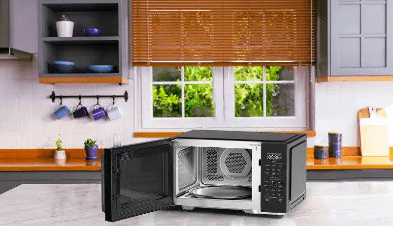 Mikrofalówka stoi otwarta na blacie w kuchni. W tle okno z żaluzjami i półki z naczyniami.