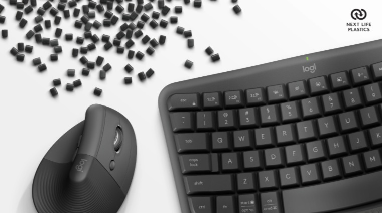 Czarna klawiatura, mysz Logitech i rozrzucone czarne granulki na białej powierzchni. Logo Next Life Plastics.