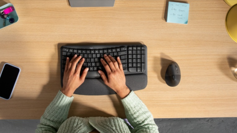 Dłonie piszące na czarnej ergonomicznej klawiaturze na drewnianym biurku, z myszą, telefonem i małymi przedmiotami.