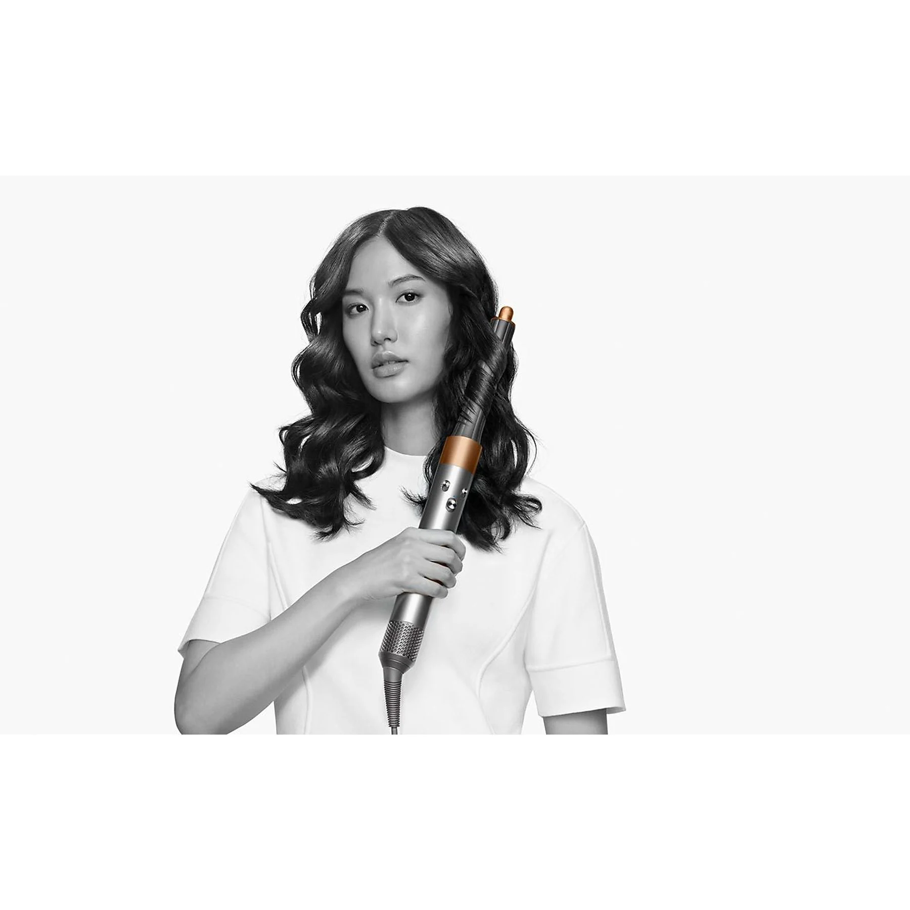 Kobieta trzyma srebrno-miedzianą lokówkę przy swoich falowanych, ciemnych włosach. Ma na sobie białą koszulę. Białe tło.