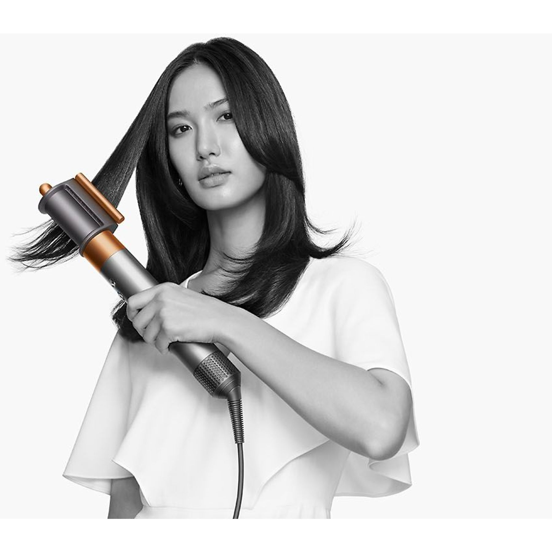 Kobieta stylizuje włosy stylizatorem Dyson. Ma na sobie białą bluzkę, tło jest białe.