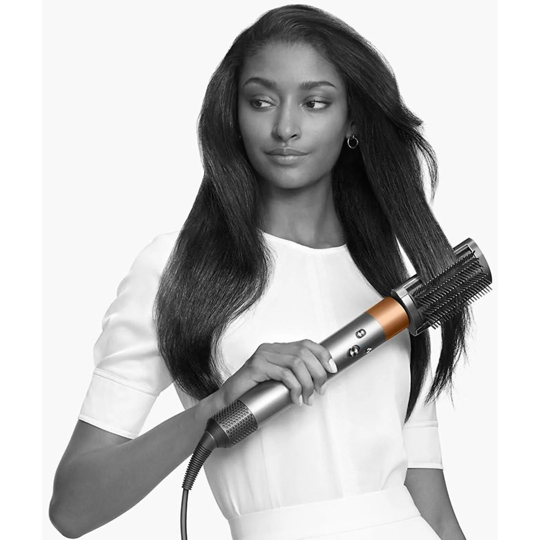 Kobieta z długimi, ciemnymi włosami stylizuje je za pomocą srebrnego i miedzianego narzędzia do stylizacji. Ma na sobie biały top.