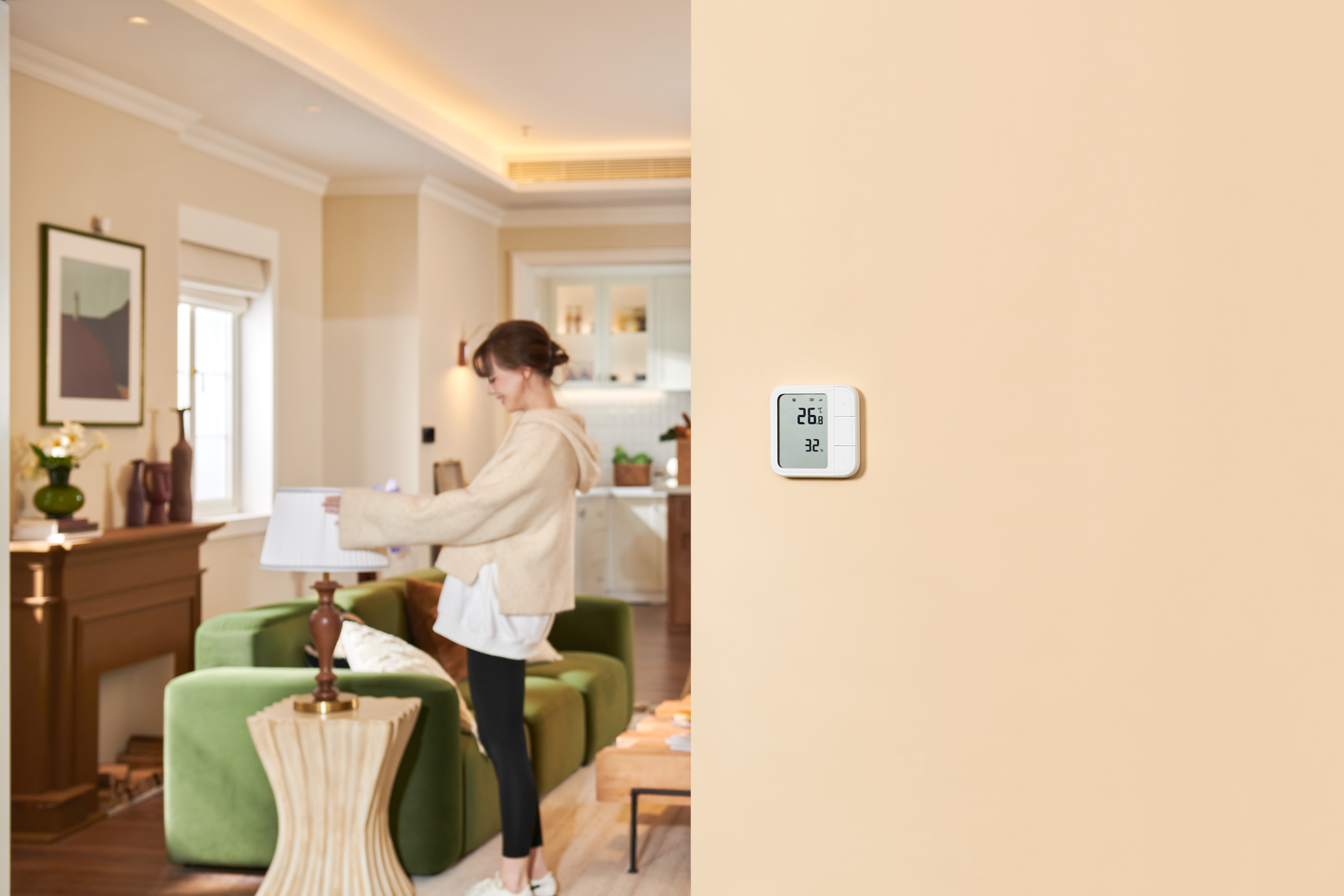 Eine Frau justiert einen Lampenschirm in einem Wohnzimmer. Ein Wandthermostat zeigt 26,8 und 32 an. Ein grünes Sofa ist sichtbar.