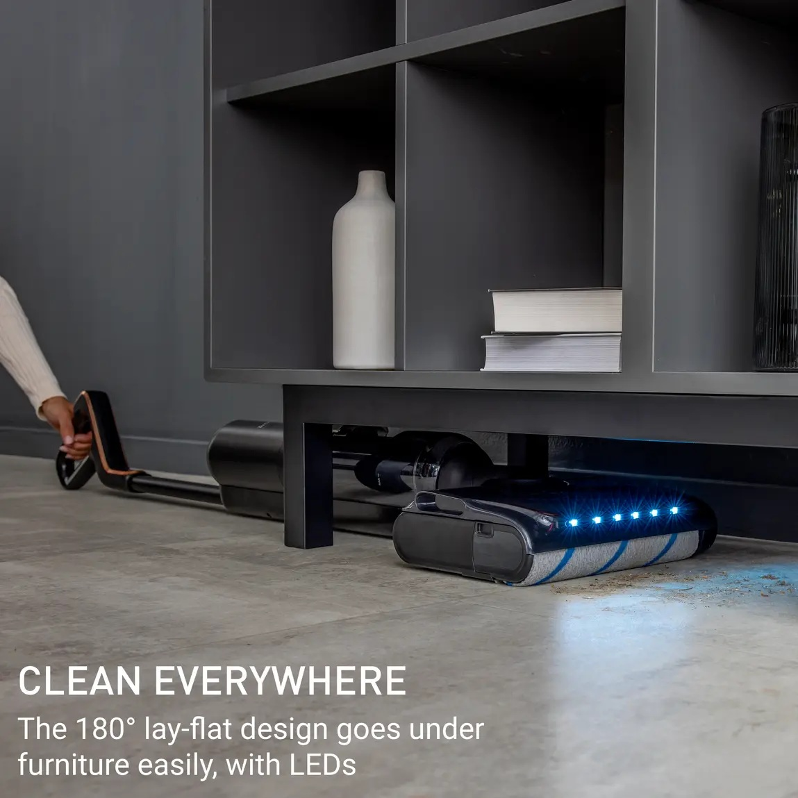 Odkurzacz znajduje się pod meblami. Ręka trzyma uchwyt. Półka z książkami i wazonem jest nad nim.