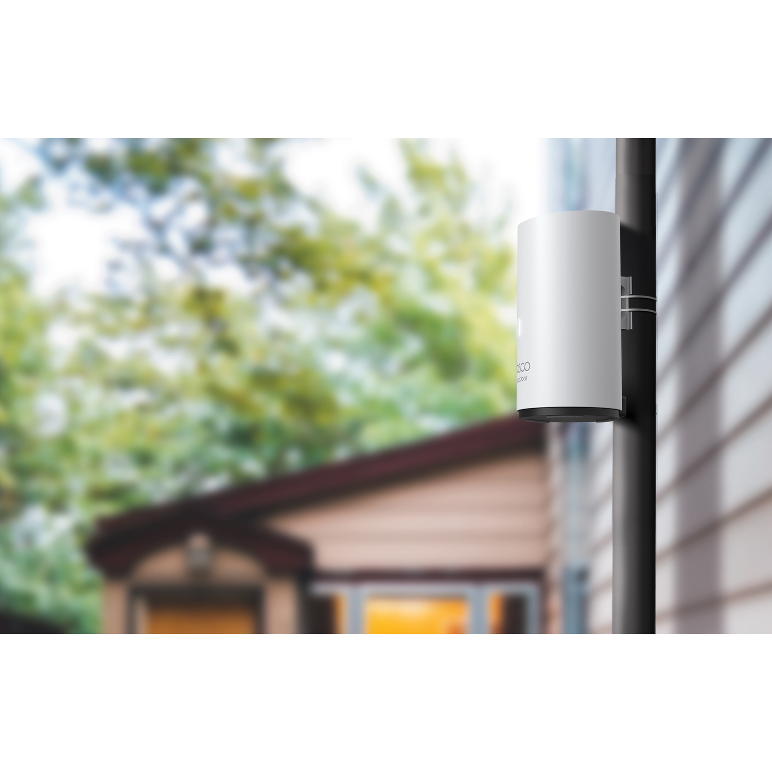 A white cylindrical device is mounted on a black pole. It's attached to a wall, with a blurred house and trees in the background.