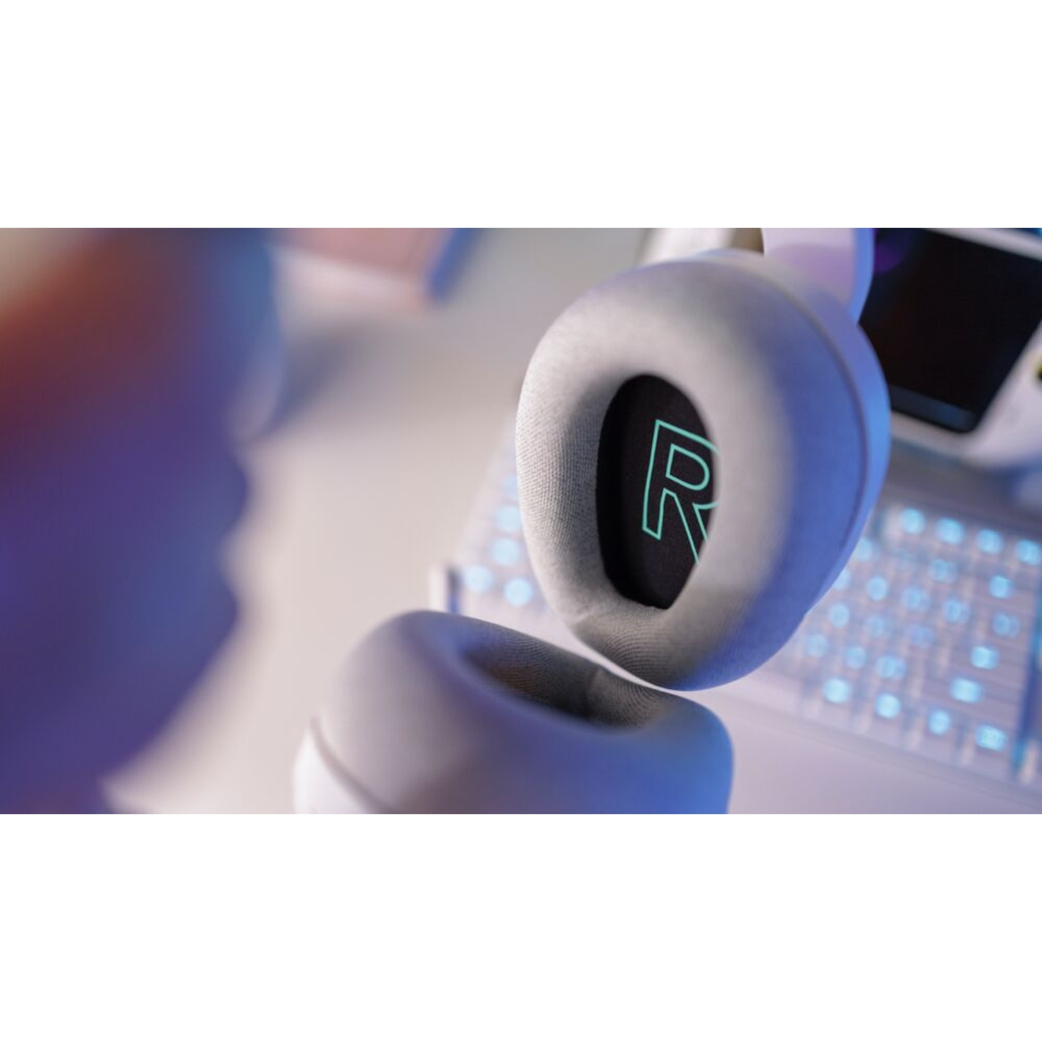Primo piano di cuffie da gioco bianche con logo 'R' illuminato, tastiera e un dispositivo portatile sullo sfondo, illuminato di blu.