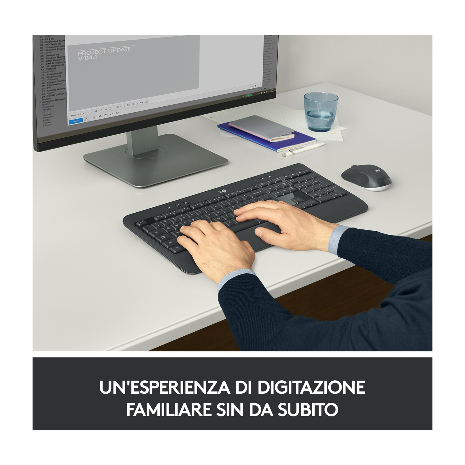 Una persona digita su una tastiera nera su una scrivania bianca. Sono visibili anche un monitor del computer, un mouse e un bicchiere d'acqua.