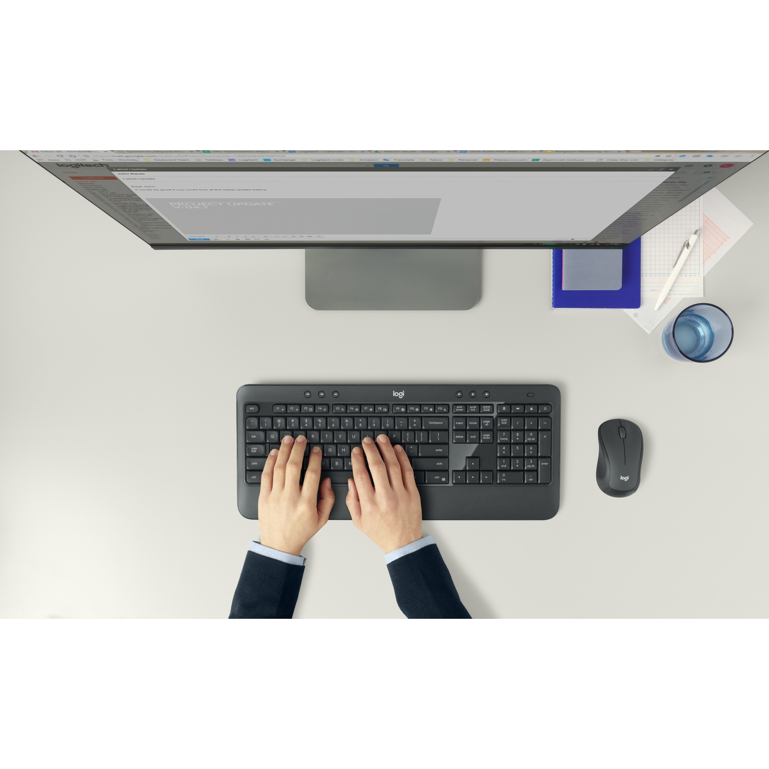 Vista dall'alto di una scrivania con tastiera, mouse, monitor e documenti. Mani che digitano sulla tastiera, un bicchiere d'acqua e una penna sono visibili.