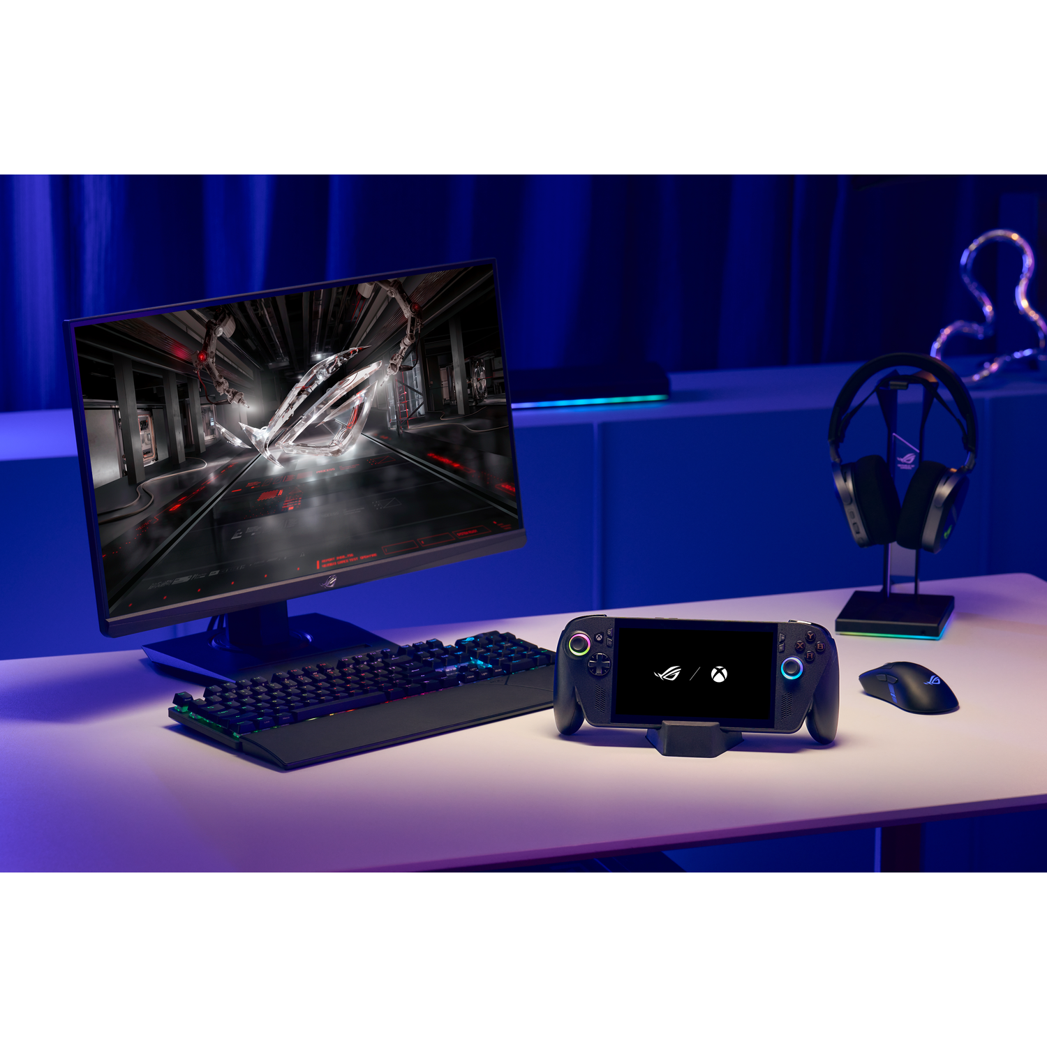 Een gaming-opstelling met een monitor met een logo, toetsenbord, handheld console, koptelefoon en muis op een witte tafel.