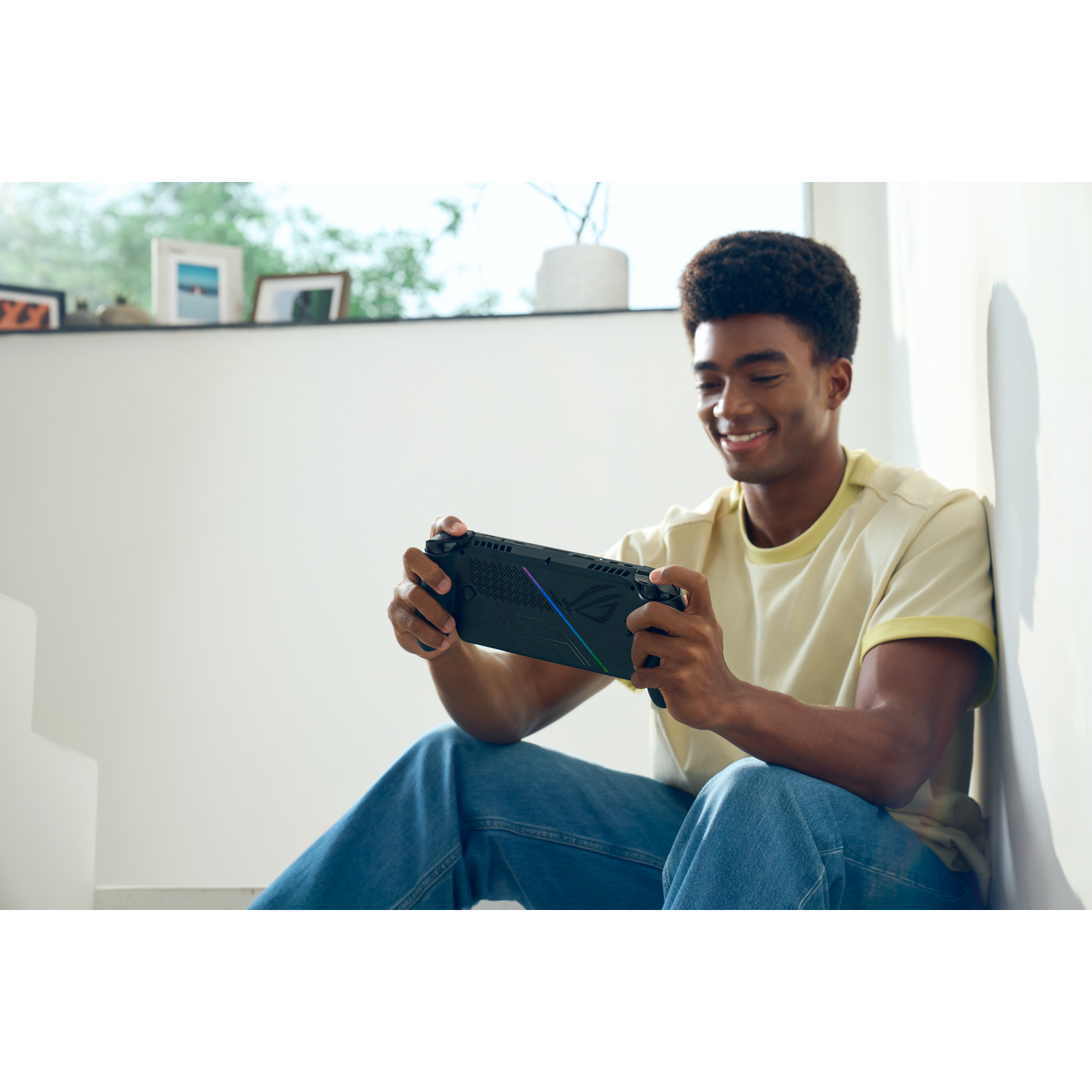 Een lachende man zit te spelen met een handheld gameconsole. Hij draagt een geel shirt en een blauwe jeans. De achtergrond toont een raam.