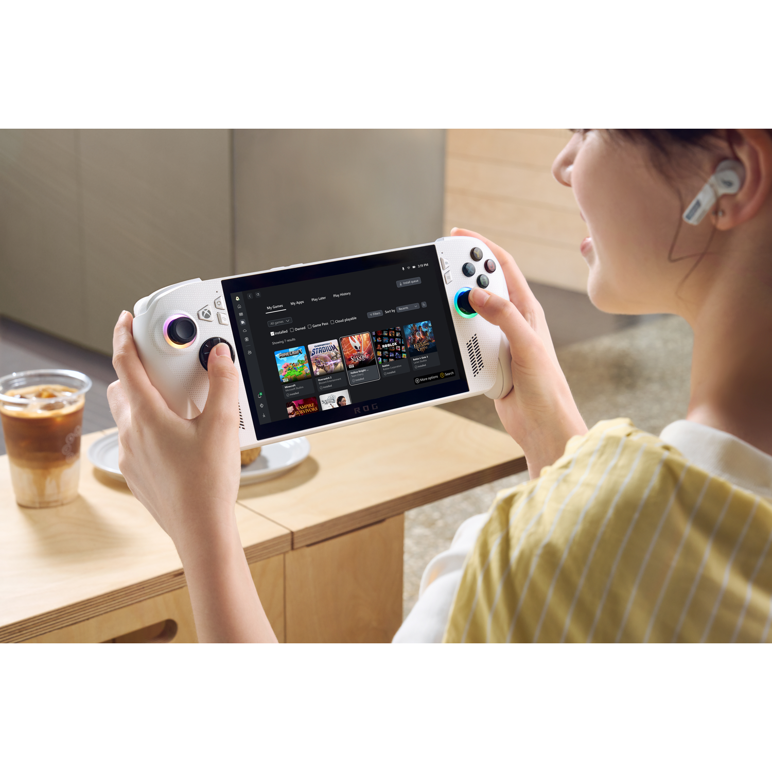 Een vrouw houdt een witte draagbare gameconsole vast, spelend. Een kopje koffie staat op tafel. Ze draagt oordopjes.