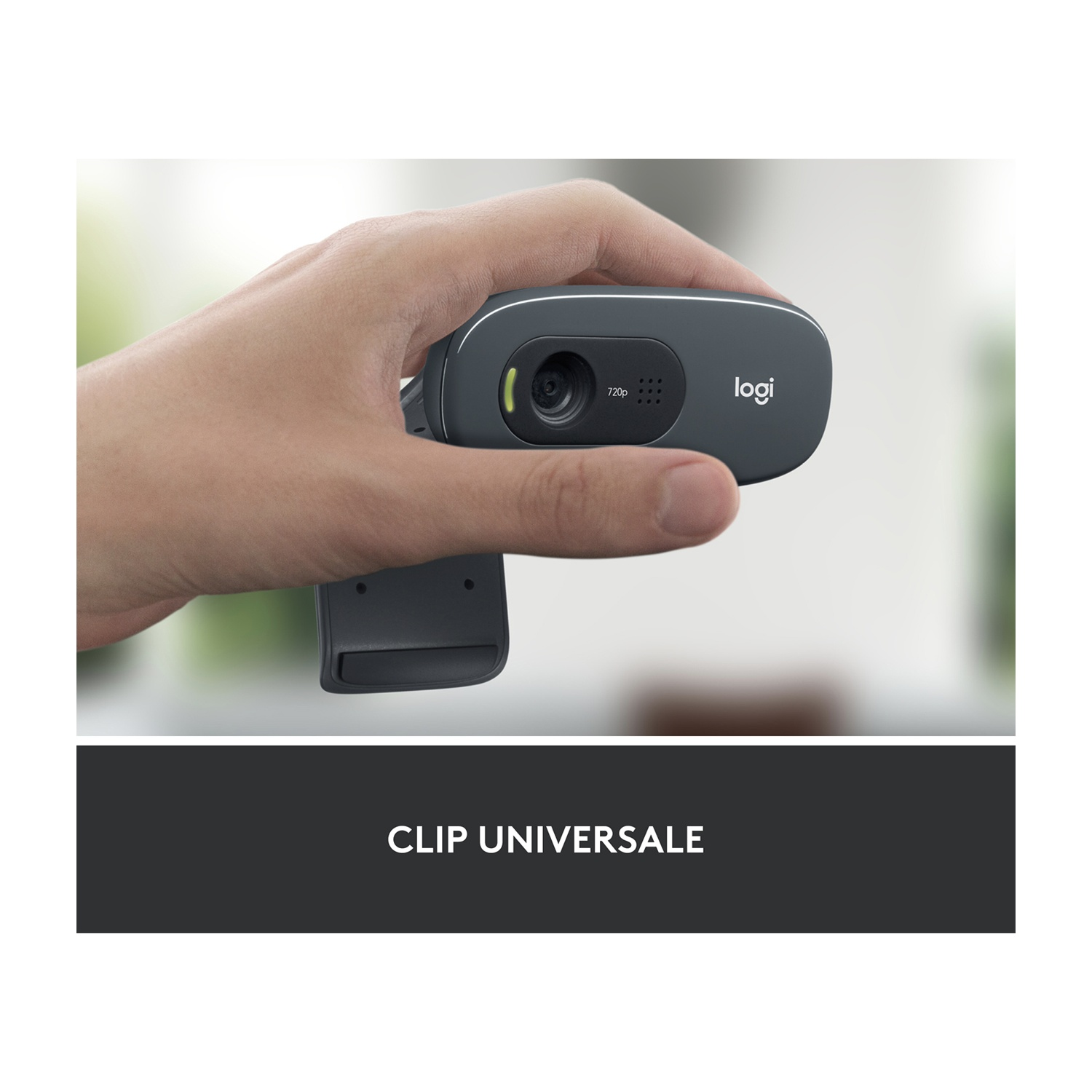 Una mano tiene una webcam Logitech grigio scuro. La webcam ha un obiettivo, 720p e il logo logi. Lo sfondo è sfocato con il verde.