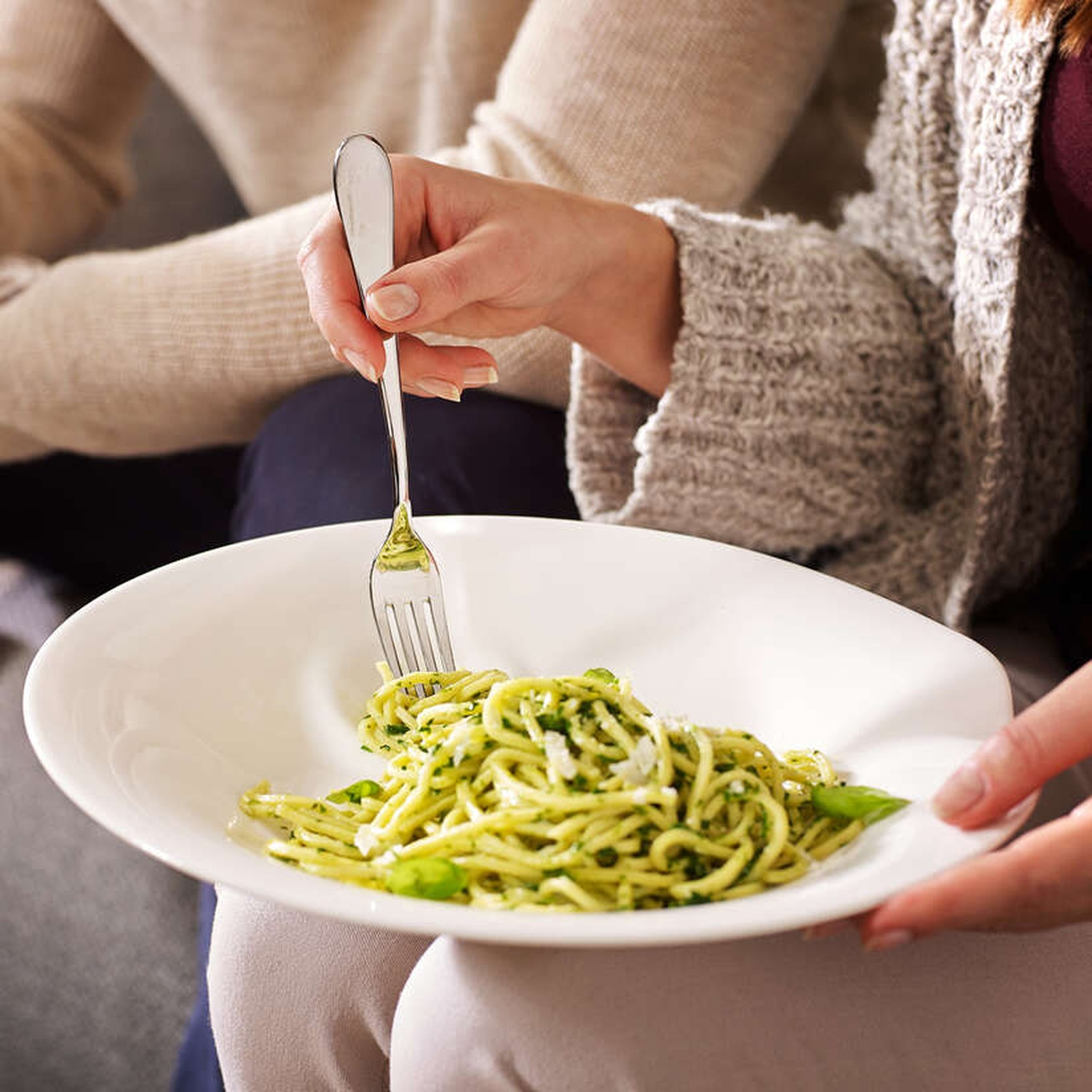 Une personne tient un bol blanc avec des pâtes au pesto. Une fourchette est dans les pâtes. Une autre personne est en arrière-plan.