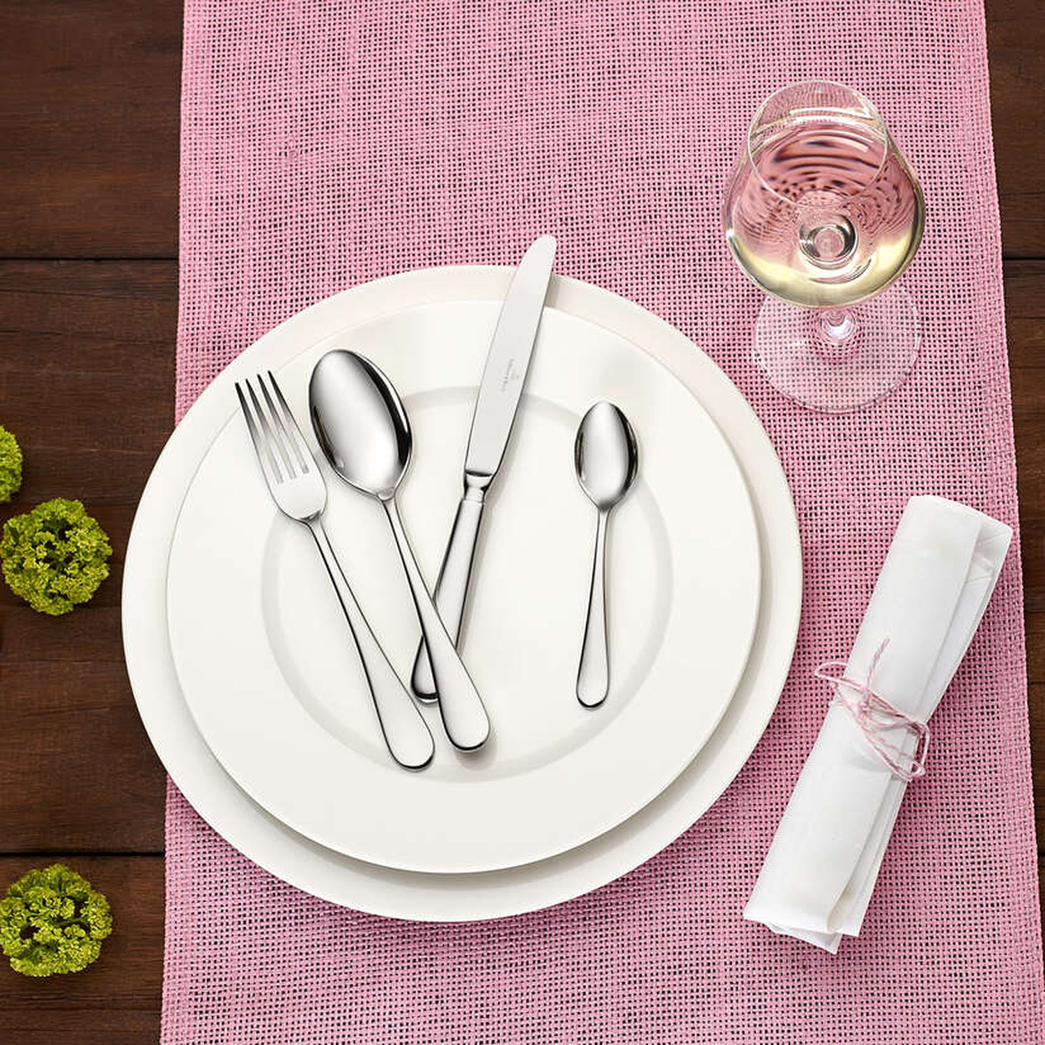 Mise en place avec assiettes blanches, couverts, verre à vin et serviette roulée sur un set de table rose, sur une table en bois.