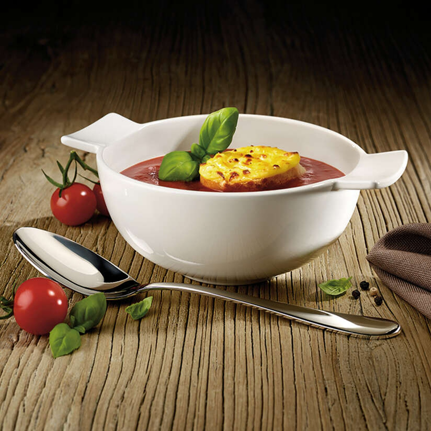 Un bol de soupe de tomates avec un croûton au fromage, du basilic et des tomates sur une table en bois avec une cuillère.