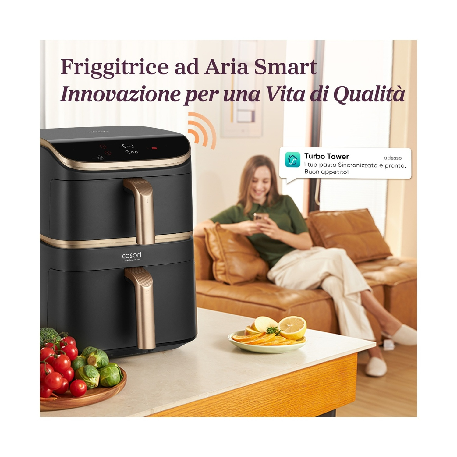 Una friggitrice ad aria con verdure e frutta su un tavolo. Una donna è seduta su un divano che usa il telefono.