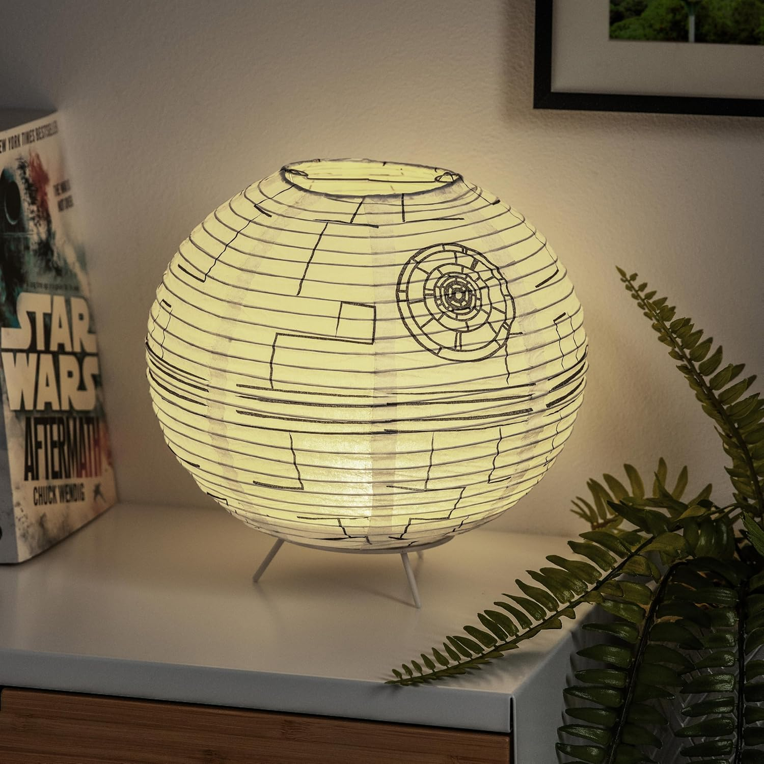 Egy Halálcsillag lámpa egy fehér felületen van, egy Star Wars könyv és egy páfrány mellett. A lámpa világít, fekete vonalak vannak rajta.