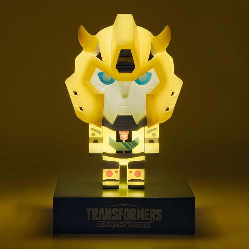 Egy sárga Bumblebee Transformer figura kék szemekkel, megvilágítva. Fekete alapon áll, rajta a Transformers Earthspark logó.