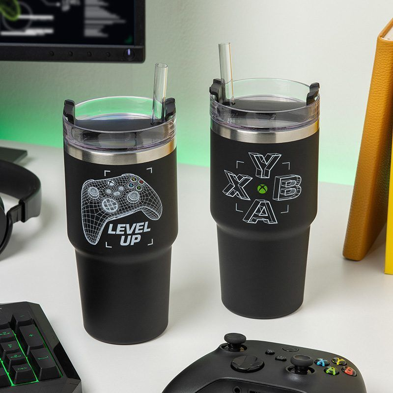 Két fekete pohár fedővel és szívószállal, az egyiken kontroller grafika és 'Level Up', a másikon 'XBA' logó. Kontrollerek az asztalon.