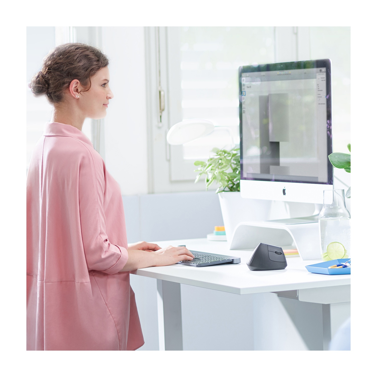 Una donna in rosa lavora a una scrivania bianca con un computer. Sono presenti una tastiera e un mouse verticale. Una finestra e delle piante sono sullo sfondo.