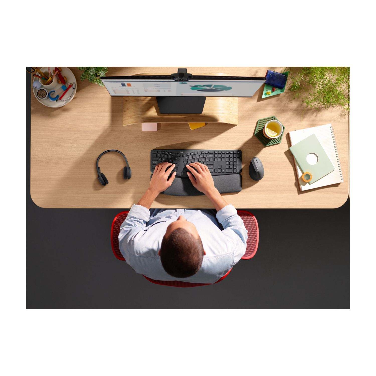 Vista dall'alto di una persona alla scrivania. Sta usando una tastiera e un mouse. Sono visibili un monitor, cuffie e altri accessori per l'ufficio.