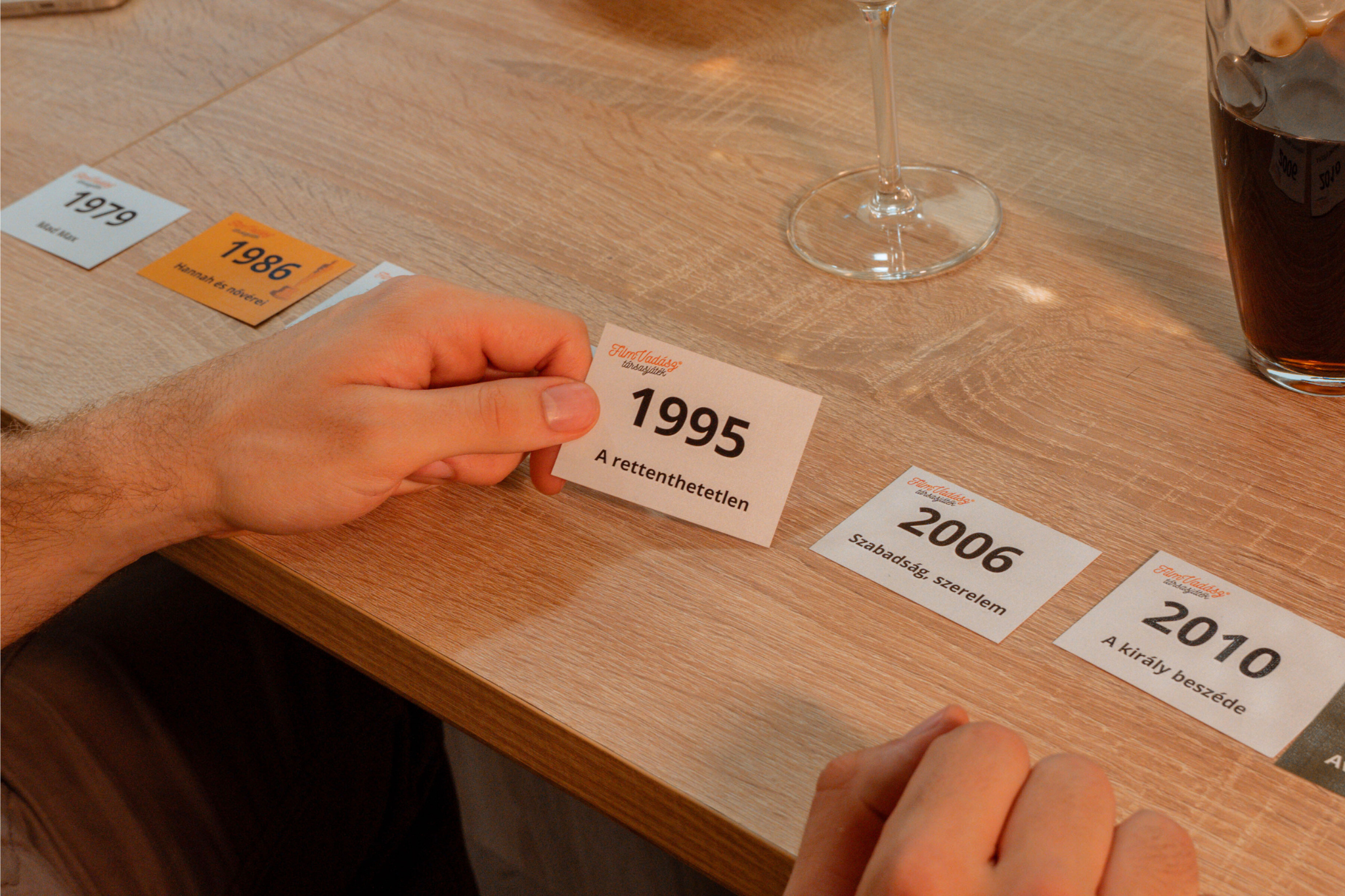 Kézben évszámokkal ellátott kártyák faasztalon, pohárral és itallal. A kártyákon 1979, 1986, 1995, 2006, 2010.
