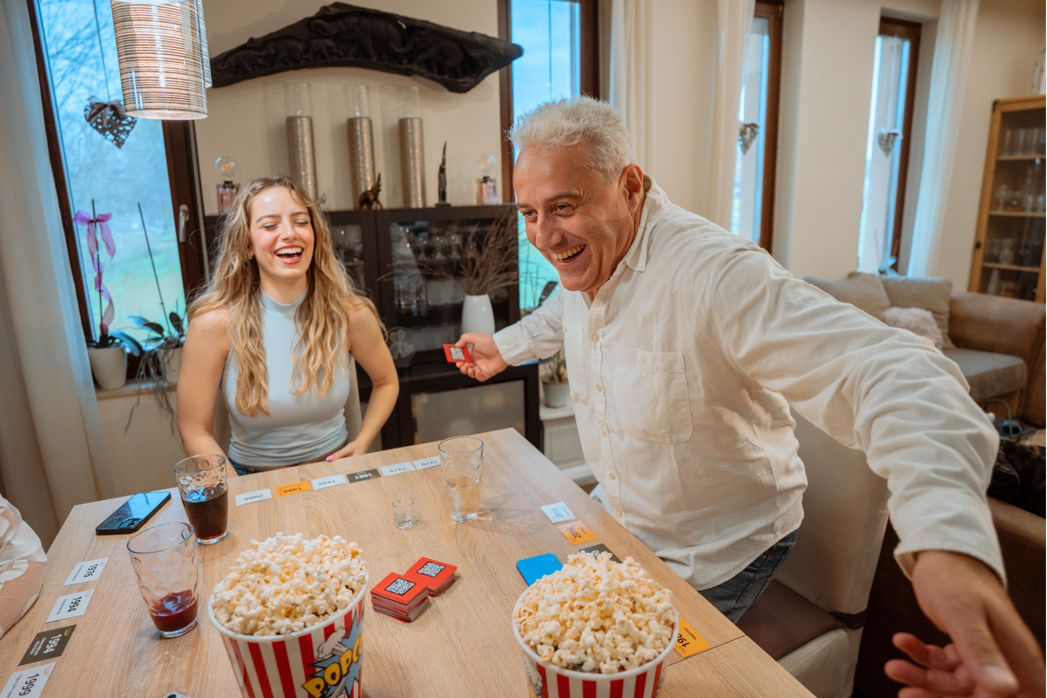 Egy férfi és egy nő nevetve játszik. Popcorn és italok az asztalon. A férfi kártyát tart.