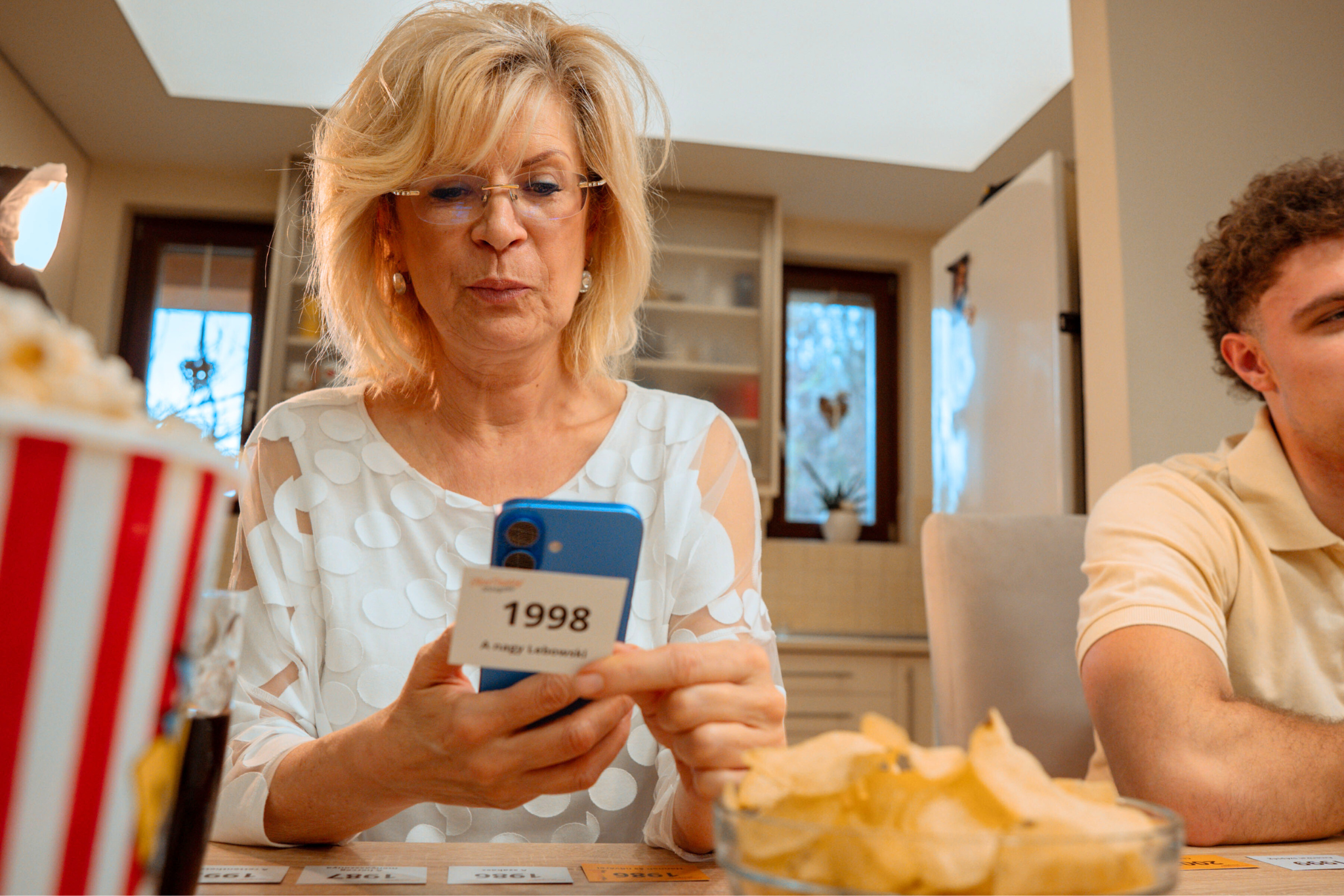 Egy nő telefont és egy '1998'-as kártyát tart. Van pattogatott kukorica, chips és egy férfi a háttérben.
